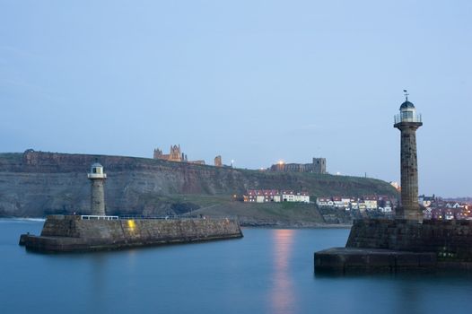 Two breakwater navigation beacons on the ends of the piers on either side of the entrance to Whitby harbour at dusk with St Marys Church and Whitby Abbey visible on Tate Hill in the distance
