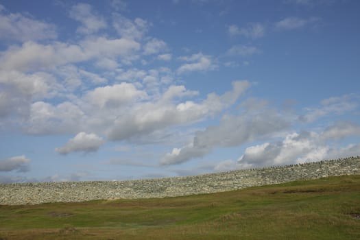 Green grass leads to an old dry stone wall built from white stone with a blue cloud sky in the distance.