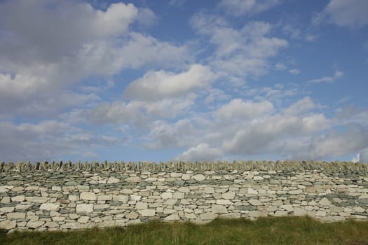 Green grass leads to an old dry stone wall built from white stone with a blue cloud sky in the distance.