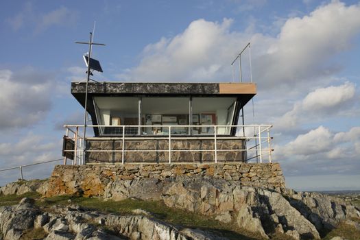 A disused coastguard station on rocks with large windows and safety rail.