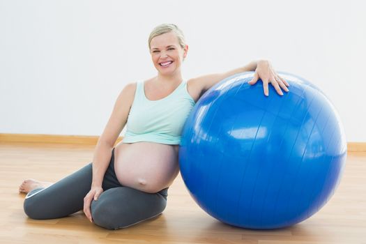 Pregnant woman sitting beside exercise ball smiling at camera in a fitness studio