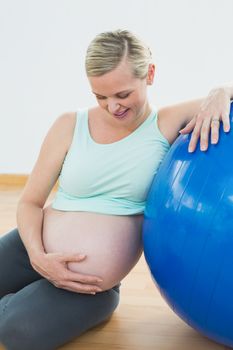 Smiling pregnant woman leaning against exercise ball holding her belly in a fitness studio