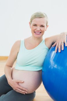 Happy pregnant woman leaning against exercise ball holding her belly in a fitness studio