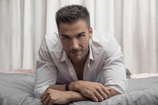 Sexy male model lying alone on his bed in his bedroom, looking at camera with a seductive attitude