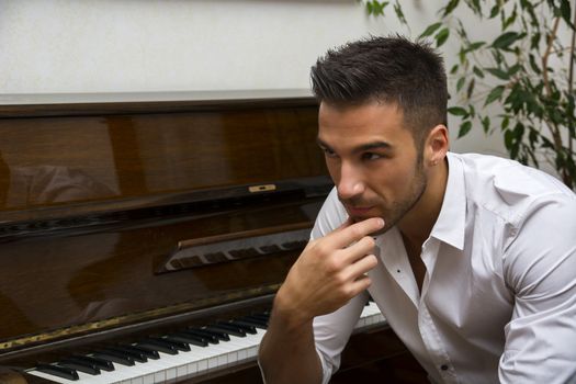Young handsome male artist smiling while sitting next to his wooden classical upright piano, indoor portrait