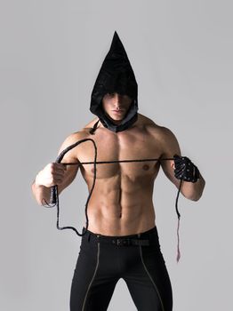 Scary muscular young man with pointed hood on naked body, holding whip in hand