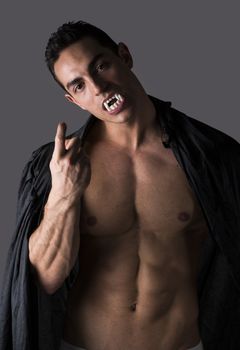 Naked muscular fit young man in briefs posing as a vampire or Dracula in a black cloak showing off his powerful body bearing his fangs with welcoming gesture