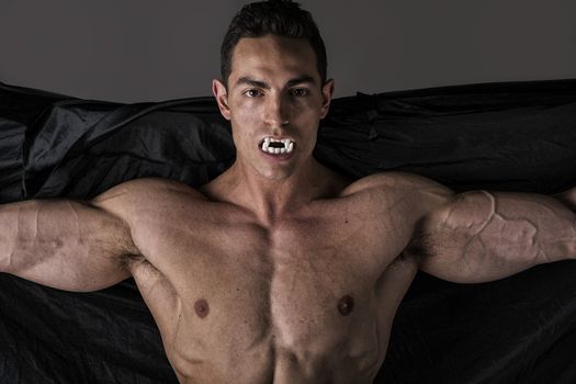 Naked muscular fit young man posing as a vampire or Dracula in a black cloak showing off his powerful body bearing his fangs with arms spread open