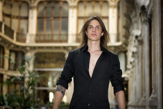 Handsome long hair man portrait in black shirt standing in elegant gallery in european city center