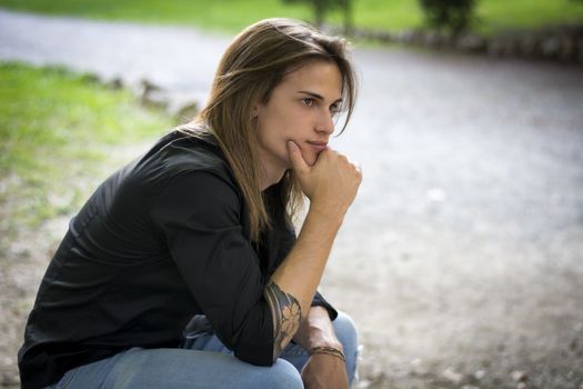 Handsome long hair young man portrait, sitting outdoor thinking, sad or worried