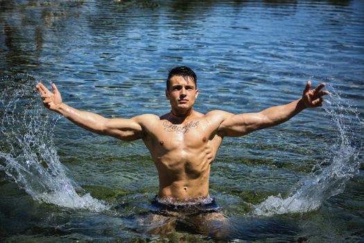 Handsome, hot young bodybuilder in the sea, splashing water up, showing his muscular torso and arms