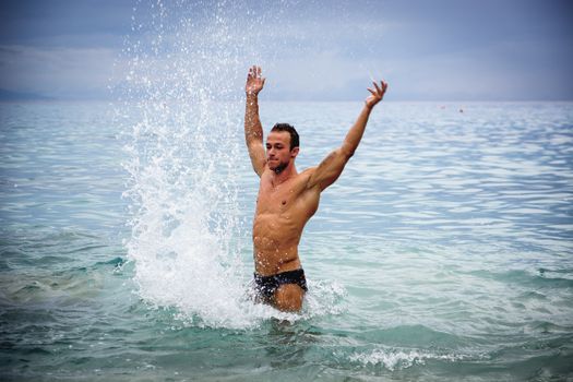 Handsome, hot young bodybuilder in the sea, splashing water up, showing his muscular torso and arms