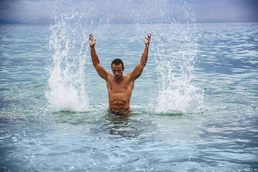 Handsome, hot young bodybuilder in the sea, splashing water up, showing his muscular torso and arms