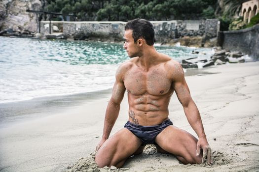 Attractive and fit young bodybuilder in bathing suit kneeling on the beach