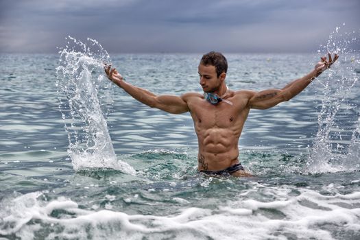 Handsome, hot young bodybuilder in the sea, splashing water up, showing his muscular torso and arms