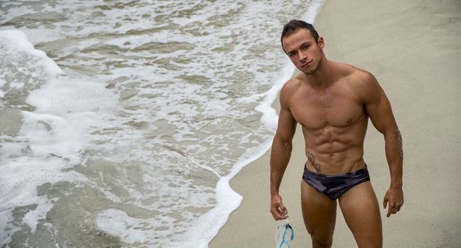 Attractive and fit young bodybuilder in bathing suit on the beach looking at camera