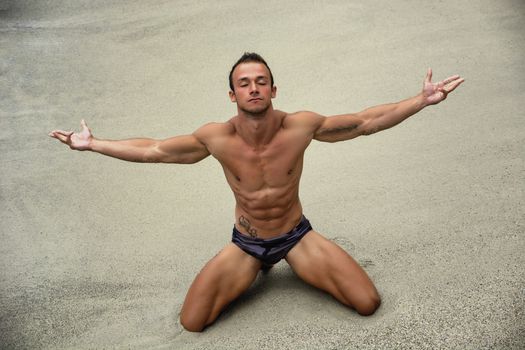 Attractive and fit young bodybuilder in bathing suit kneeling on the beach with arms spread open and eyes closed