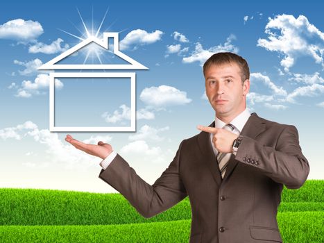 Businessman in a suit hold house icon on background of green grass and blue sky