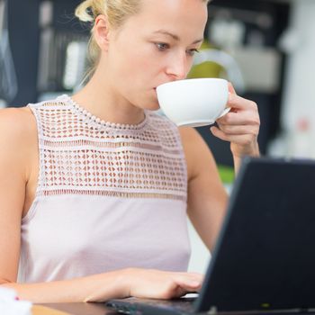 Business woman working remotly from her dining table while having her morning coffee. Home kitchen in the background.