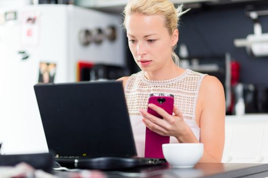 Business woman working from her home. Multitasking. Having morning coffee while texting on the smarth phone and working on leptop computer.