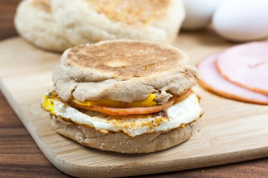 English muffin, egg, ham, and cheese breakfast sandwich on a cutting board with ingredients in the background.