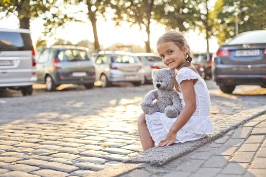 Small girl sitting near city road with her toy teddy bear