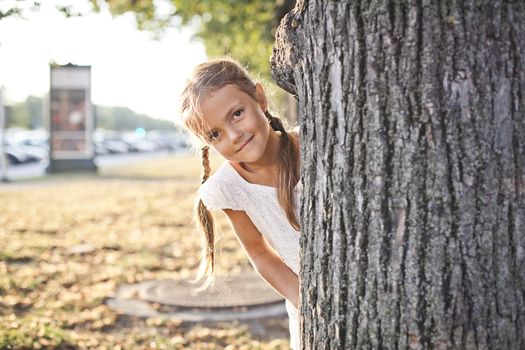 Young girl is hiding behind the tree in a city park