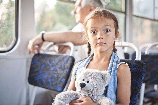 Small surprised girl sit in a tram with her toy teddy bear
