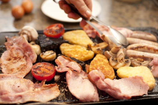 Man serving a hearty cooked breakfast warming on the griddle with bacon, tomato, hash browns, mushrooms and sausages