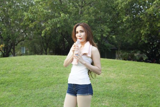 Female breaks standing and holding a bottle of water. On the grass in the public park. While jogging