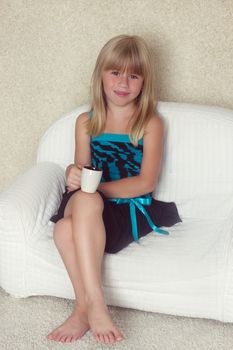 Girl 5 years old sitting on a sofa in a dress with a cup in hands