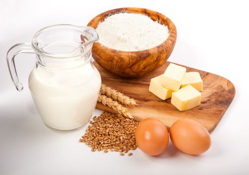 Glass jug with milk, wheat seeds, flour and two eggs on white background