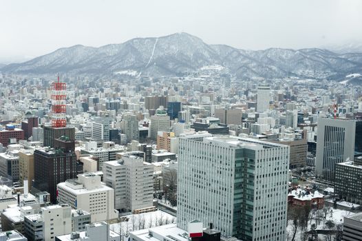 City of Sapporo in Japan as viewed from the JR Tower with snow on the buildings and a panoramic view showing the topography of the surrounding mountains behind the modern architecture