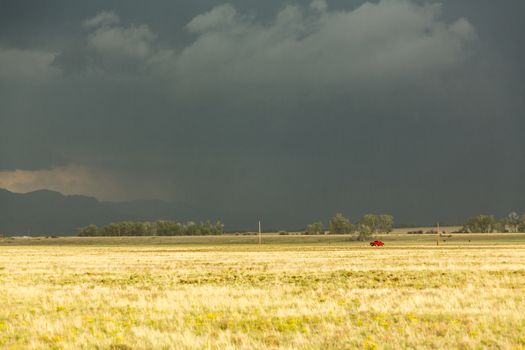 Sun illuminating prairie grassland with dark and forbidding thunderclouds and downpour with red van riding along lonely country road