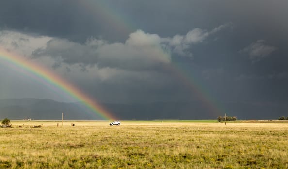 Sun illuminating prairie grassland with dark and forbidding thunderclouds and downpour with van riding along lonely country road and rainbow ending over the vehicle
