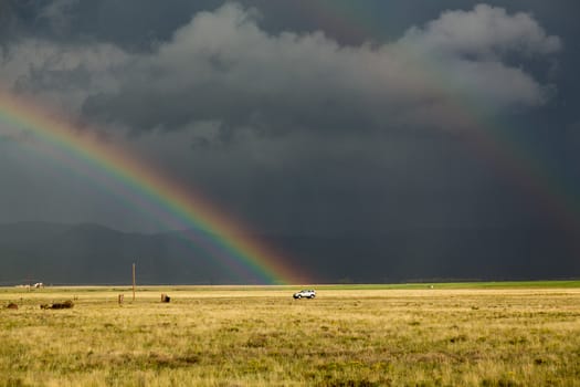 Sun illuminating prairie grassland with dark and forbidding thunderclouds and downpour with van riding along lonely country road and rainbow ending over the vehicle