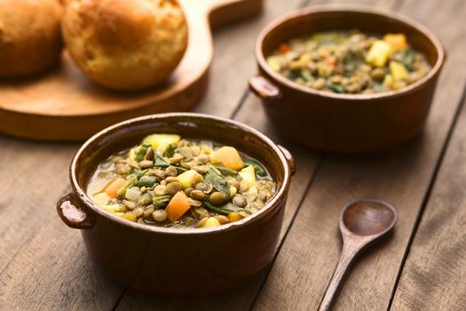 Vegetarian soup made of lentils, spinach, potato, carrot and onion served in dark brown bowls with buns in the back (Selective Focus, Focus in the middle of the soup)