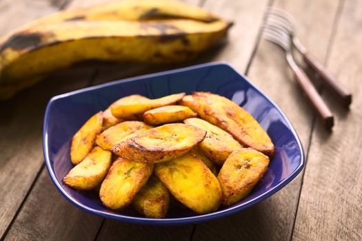 Fried slices of the ripe plantain in blue bowl, which can be eaten as snack or is used to accompany dishes in some South American countries, ripe plantains in the back (Selective Focus, Focus on the front of the upper plantain slice)