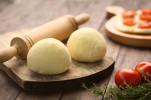 Small balls of yeast dough for pizza or flatbread with rolling pin on wooden board, tomato and thyme in the front, tomato flatbread in the back (Selective Focus, Focus on the front of the first dough ball)