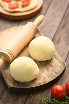 Small balls of yeast dough for pizza or flatbread with rolling pin on wooden board, tomato and thyme in the front, tomato flatbread in the back (Selective Focus, Focus on the front of the first dough ball)