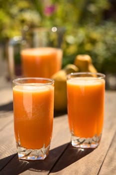 Two glasses of freshly prepared papaya juice with pitcher and papaya fruits in the back on table outdoors (Selective Focus, Focus on the front rim of the first glass) 