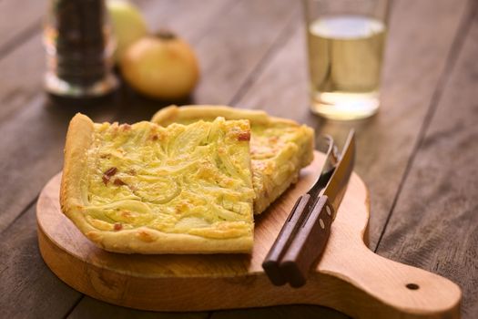 Pieces of traditional German Zwiebelkuchen or onion cake, made of a yeast dough and topped with onions and bacon in cream sauce with cutlery on wooden board, white wine in the back (Selective Focus, Focus in the middle of the first piece) 