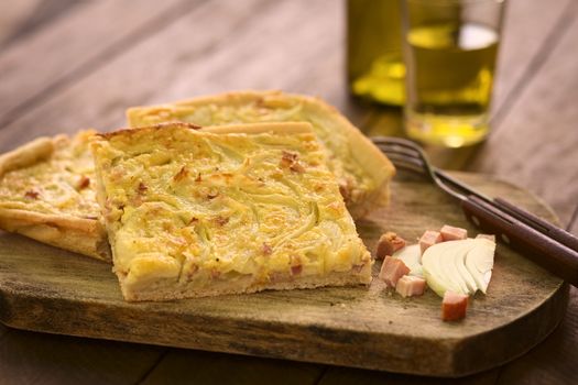 Pieces of traditional German Zwiebelkuchen or onion cake, made of a yeast dough and topped with onions and bacon in cream sauce with cutlery and ingredients on wooden board, white wine in the back (Selective Focus, Focus one third into the first piece) 