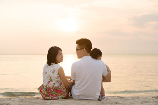 Portrait of young Asian family seated on beach outdoor vacation, during summer sunset, natural sunlight. 
