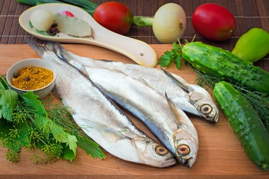 On a table on a chopping board there are a fish, cucumbers, tomatoes, onions, spices and parsley greens.
