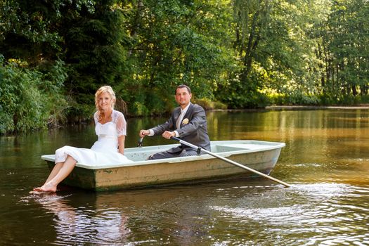 beautiful young wedding couple, blonde bride with flower and her groom just married on small boat at pond with evening sun