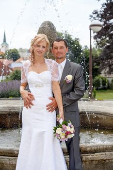 beautiful young wedding couple, blonde bride with flower and her groom against fountain