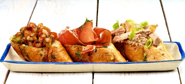 Tapas Bruschetta with Bacon and Vegetables, Cured Ham and Pate with Spring Onion on Garlic Bread closeup on Plate on White Wooden background