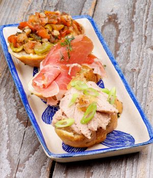 Tapas Bruschetta with Pate with Spring Onion, Cured Ham and Bacon with Vegetables on Garlic Bread on Plate isolated on Rustic Wooden background