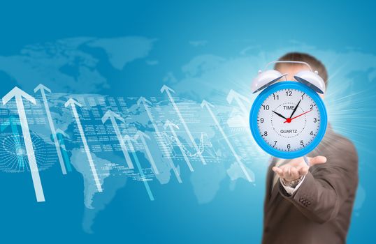 Business man hold alarm clock in hand. World map, graphs and arrows as backdrop